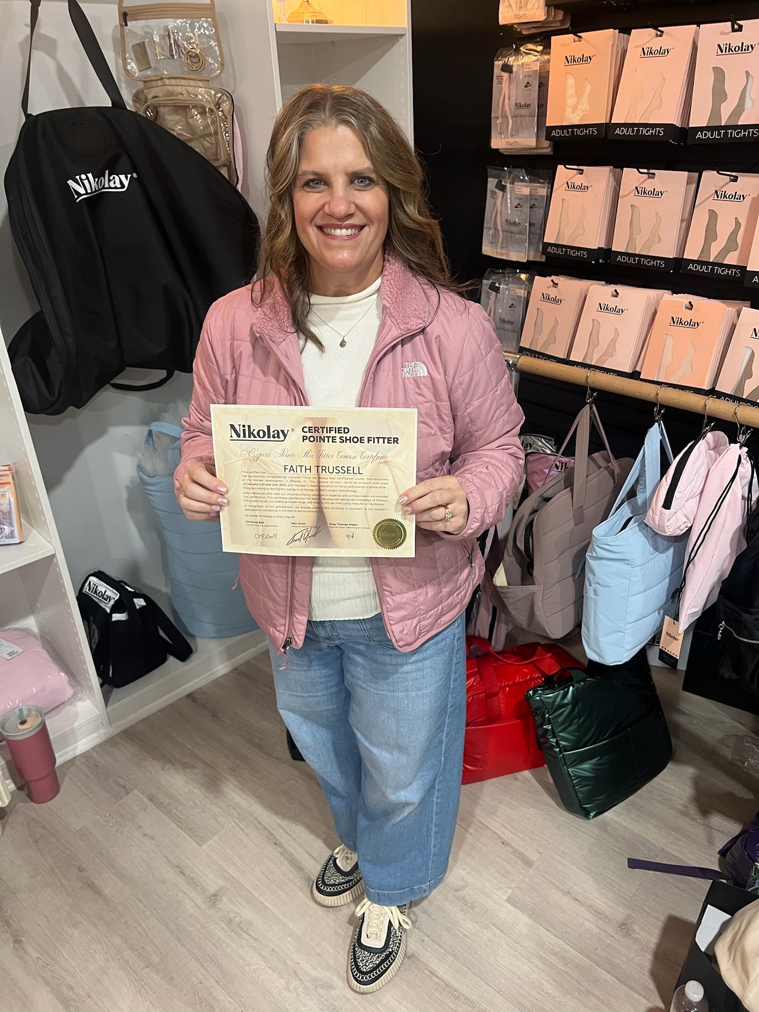 Faith from Dance Fantasy stands in a retail store holding a Nikolay 'Certified Authenticity Letter'. She is wearing a pink puffer jacket, white t-shirt, blue jeans, and black-and-white sneakers.